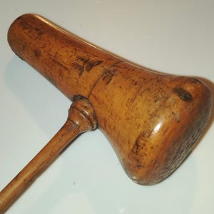 Könnte beinhalten: Ein Holzhammer mit einem zylindrischen Kopf und einem langen, schlanken Stiel. Das Holz hat eine natürliche hellbraune Oberfläche mit sichtbarer Maserung, oberflächlichen Kratzern und Gebrauchsspuren am gesamten Werkzeug.