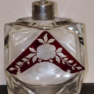 Könnte beinhalten: Ein quadratischer Parfümflakon aus klarem Glas mit facettierten Kanten. Die Vorderseite zeigt eine dreieckige, tiefrote Auflage mit einem eingravierten weißen Rosenmotiv. Er hat einen strukturierten, silberfarbenen Metalldeckel mit einer kleinen Düse.