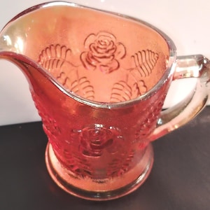Pode incluir: Uma pequena jarra de vidro vermelho iridescente com um padrão em relevo de rosas florescentes e folhas. O recipiente possui um bico largo, uma alça lateral resistente e uma base circular, com um acabamento metálico cintilante.