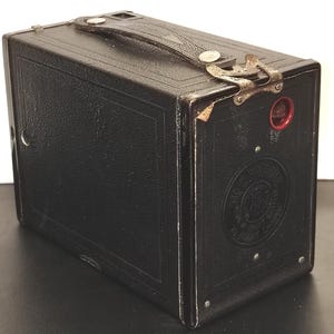 May include: A black box camera with a textured finish, featuring a top carrying handle, a metal latch, and a circular red viewing window. The side panel displays an embossed circular logo with the text No. 2 Brownie and Kodak Limited.