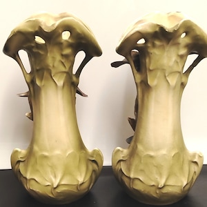 Può includere: Una coppia di vasi in ceramica abbinati con una finitura sfumata in verde pallido e crema. Ogni vaso presenta un bordo svasato e smerlato con dettagli intagliati e un motivo organico a foglie in rilievo modellato sulla base e sul collo.