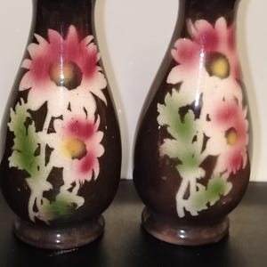 May include: A pair of dark brown ceramic vases with a glossy finish, each featuring a hand-painted floral design of pink and white flowers with yellow centres and green leaves. The vases have a narrow neck, a flared base, and a metallic rim.
