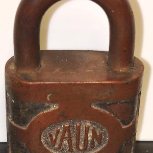 Pode incluir: Um cadeado de metal castanho-escuro desgastado com um arco arredondado e um corpo retangular. A parte frontal do cadeado apresenta um emblema oval central com um fundo texturizado de cor clara contendo as letras maiúsculas em relevo VAUN.