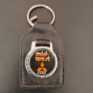 Könnte beinhalten: Ein schwarzer Lederschlüsselanhänger mit einem kreisförmigen, silberfarbenen Metallemblem mit dekorativem Rand. Das Emblem zeigt den orangefarbenen Schriftzug mid-west, ein rotes Mitsubishi-Logo und den weißen Schriftzug COLT. Ein silberner Metallring ist oben befestigt.
