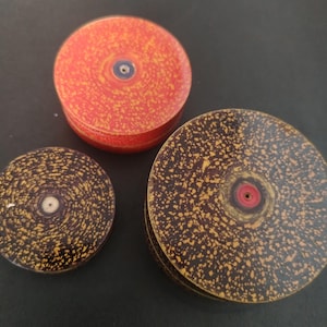 Op de afbeelding: Drie cirkelvormige objecten met gespikkelde patronen. Eén is felrood met gele spikkels en een donker centrum. Twee zijn donkerbruin met gele spikkels, voorzien van een centraal gat met een rode of witte ring. Deze items hebben een platte schijfvorm.