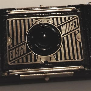 May include: A black Ensign Midget Model 22 camera featuring a rectangular silver metal faceplate with vertical black stripes, a central circular lens housing, and engraved text reading Ensign Midget Model 22 on the front panel.