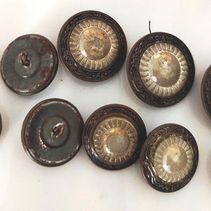 Può includere: Una collezione di dieci bottoni rotondi caratterizzati da un bordo esterno in metallo marrone scuro con un intricato motivo in rilievo e un inserto circolare centrale con una finitura riflettente, chiara e leggermente iridescente con creste radiali.