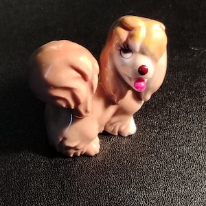 May include: A small tan figurine of a dog with long, flowing ears, a white muzzle, a dark red nose, and a bright pink tongue. The dog has painted black eyelashes and a large, rounded tail, rendered in a smooth, glossy finish with light brown highlights.