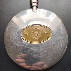 Könnte beinhalten: Ein runder, silberfarbener Metallflachmann mit Schraubverschluss. In der Mitte befindet sich eine ovale, messingfarbene Plakette mit dem geprägten Text "SCHWEPPES SINCE 1783", der eine Brunnen-Grafik umgibt, flankiert von zwei kleinen Pfeilsymbolen.