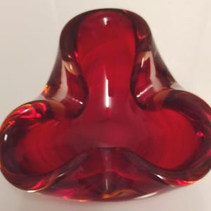 May include: A deep red glass ashtray with a distinct three-lobed, clover-like shape. The thick, translucent glass features smooth, rounded edges and a central basin, creating a reflective surface that catches light throughout the curved form.