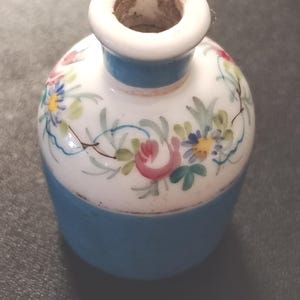 Op de afbeelding: Een kleine keramische vaas met een rond lichaam en een smalle hals. Het onderste gedeelte is effen lichtblauw, terwijl het bovenste gedeelte wit is en versierd met een handgeschilderd bloemmotief met roze rozen, blauwe madeliefjes en groene bladeren.