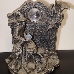 May include: A silver-coloured figurine depicting a robed wizard standing at a table with a small glowing orb, accompanied by a dragon perched on a stone archway. The structure features intricate relief carvings and two embedded clear faceted crystals.