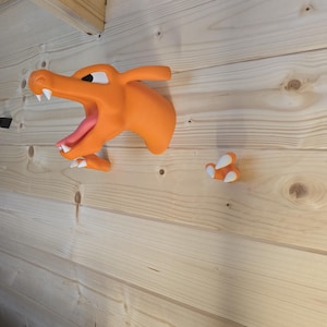 May include: A wall-mounted orange dragon head sculpture with an open mouth, white teeth, and a pink tongue, accompanied by a separate orange claw piece with white nails, both featuring a smooth matte finish and mounted on a wooden surface.