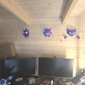 May include: Three purple wall-mounted figures depicting cartoon creatures, including one with spiky hair, one with pointed ears, and one with claws. Below, a black gaming console, a grey controller, and two white and blue figurines are on a desk.