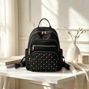May include: A black backpack featuring a quilted front pocket adorned with small silver studs. The bag includes a top handle, adjustable shoulder straps, and multiple zippered compartments with gold-toned hardware, providing a structured design.