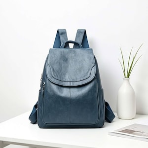 May include: A blue backpack with a smooth, slightly textured finish, featuring a top flap closure, a central vertical seam, and adjustable shoulder straps. The bag includes side pockets and a top handle, showcasing a structured design with silver-toned metal zippers on the side compartments.