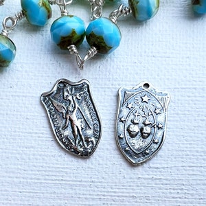 May include: A silver rosary with blue and green glass beads and two silver charms. One charm features a winged angel and the other charm features a cross and two hearts.