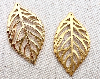 May include: Two gold-colored leaf-shaped charms. Each charm has a detailed, openwork design with veins and a small hole at the top for stringing. The charms are set against a textured, off-white background.