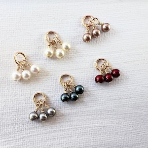 Könnte beinhalten: Sechs Sätze mit jeweils drei Perlenanhängern, jeder Satz mit einer anderen Perlenfarbe: weiß, grau, schwarz, rot, rosa und beige. Jeder Satz ist an einer goldfarbenen Schlaufe befestigt.