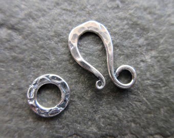 Könnte beinhalten: Ein silberfarbenes Schmuckverschlusset, bestehend aus einem gehämmerten Haken und einem strukturierten kreisförmigen Ring. Der Verschluss ist für einen sicheren Verschluss in Halsketten, Armbändern oder anderen Schmuckkreationen konzipiert. Das Metall hat ein glänzendes Finish.