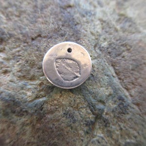 May include: A silver charm with a leaf design. The charm is round and has a small hole at the top for attaching to a necklace or bracelet.