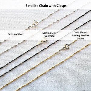 May include: Four satellite chains with clasps. The chains are silver, gunmetal, gold plated, and a two-tone gold and silver. The chains are laid out on a white surface.