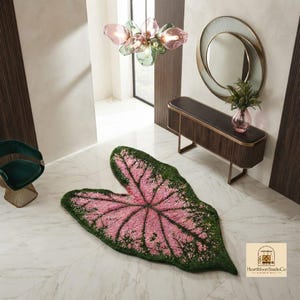 May include: A large, leaf-shaped rug featuring a vibrant pink centre with dark green edges, mimicking the appearance of a caladium leaf. The rug is placed on a white marble floor in a room with a dark wood console table and a glass chandelier.