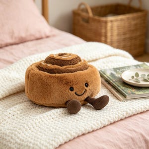 Peut inclure: Une peluche en forme de roulé à la cannelle avec une fourrure marron douce, un dessus en spirale et un visage souriant aux yeux noirs. Le jouet présente deux petites jambes en velours côtelé marron foncé à l'avant, offrant un aspect tactile.