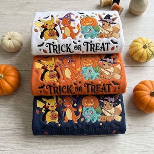 Op de afbeelding: Drie gevouwen sweatshirts in wit, oranje en marineblauw, elk met een grafische print van Pikachu, Charmander, Bulbasaur en Eevee in Halloween-kostuums met pompoenen en de tekst Trick or Treat in een zwart cursief lettertype.