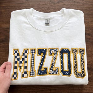 May include: A white crewneck sweatshirt featuring the word MIZZOU in large, block-style letters. Each letter has a gold embroidered border and a unique pattern, including checkerboard, gingham, stars, daisies, polka dots, and leopard print.
