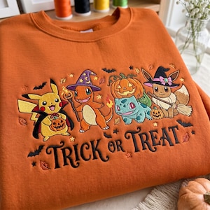 Könnte beinhalten: Ein orangefarbenes Sweatshirt mit einer Stickerei von vier Zeichentrickfiguren in Halloween-Kostümen, inklusive Kürbis, Fledermäusen und Blättern. Der Text "TRICK or TREAT" ist in schwarzer Schreibschrift unter den Figuren eingestickt.