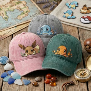 May include: Three baseball caps in pink, gray, and green, each featuring an embroidered character design. The pink cap displays Eevee, the gray cap shows Squirtle with water accents, and the green cap depicts Charmander with flames.