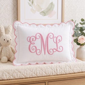 May include: A rectangular white pillow featuring a scalloped edge trimmed in pink piping. The center of the pillow displays a large, decorative embroidered monogram in pink thread with the letters E, M, and L in a cursive, swirling script style.