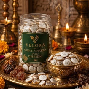 Puede incluir: Un frasco de plástico transparente lleno de conchas Gomti Chakra en espiral blancas y marrones, con una etiqueta verde con el texto VELORA GOMTI CHAKRA SACRED OFFERING. Un pequeño cuenco de latón y una bandeja dorada contienen conchas adicionales.