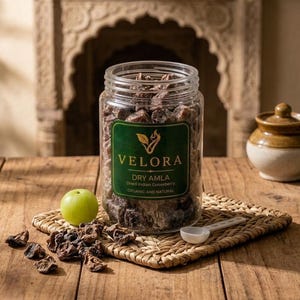 Könnte beinhalten: Ein durchsichtiges Kunststoffglas gefüllt mit dunkelbraunen getrockneten Amla-Stücken, mit einem grünen Etikett mit dem Text VELORA DRY AMLA Dried Indian Gooseberry ORGANIC AND NATURAL. Eine frische grüne Stachelbeere und lose Stücke.