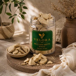 May include: A clear plastic jar filled with beige Multani Mitti clay chunks, featuring a dark green label with the text VELORA MULTANI MITTI WHOLE PURE AND NATURAL. A small ceramic bowl contains additional clay pieces, with more scattered on the surface.
