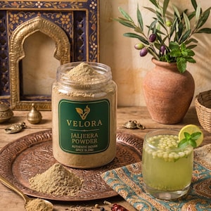 May include: A clear plastic jar of Velora Jaljeera Powder, an authentic Indian spice blend, sits on a copper tray with a pile of the light brown powder, a spoon, cumin seeds, peppercorns, a chilli, and a glass of green drink with small beads.