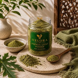 Pode incluir: Um frasco de plástico transparente cheio de pó de folha de papaia verde, com um rótulo verde escuro com o texto VELORA PAPAYA LEAF POWDER PURE AND NATURAL. O frasco assenta sobre um tabuleiro de pedra redondo com uma colher de madeira.