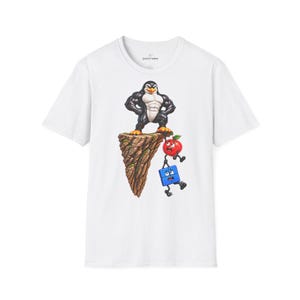 Può includere: Una t-shirt bianca a maniche corte con la grafica di un pinguino muscoloso in piedi su una scogliera rocciosa, che tiene una mela rossa per il picciolo, la quale a sua volta tiene per mano un personaggio quadrato blu. Il marchio jostromo è stampato.