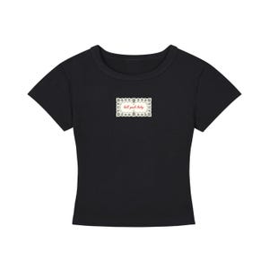 Puede incluir: Una camiseta negra de manga corta con cuello redondo. Centrado en el pecho hay un gráfico rectangular con un borde blanco decorativo de patrones intrincados y el texto "hell yeah baby" escrito en una fuente cursiva de color rojo.