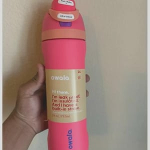 Op de afbeelding: Een felroze Owala waterfles met een beige label met de tekst: Hi there. I'm leak proof. I'm insulated. And I have a built-in straw. 24 oz./710ml. De dop is roze en perzikkleurig met een drukknop en een hendel om te ontgrendelen.