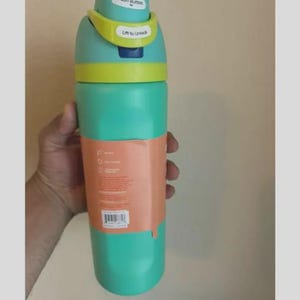 Op de afbeelding: Een talingkleurige waterfles met een limoengroen en talingkleurig deksel met een drukknop en een ontgrendelingsmechanisme. De fles heeft een perzikkleurig papieren etiket rond het midden met gedrukte tekst, iconen en een streepjescode.