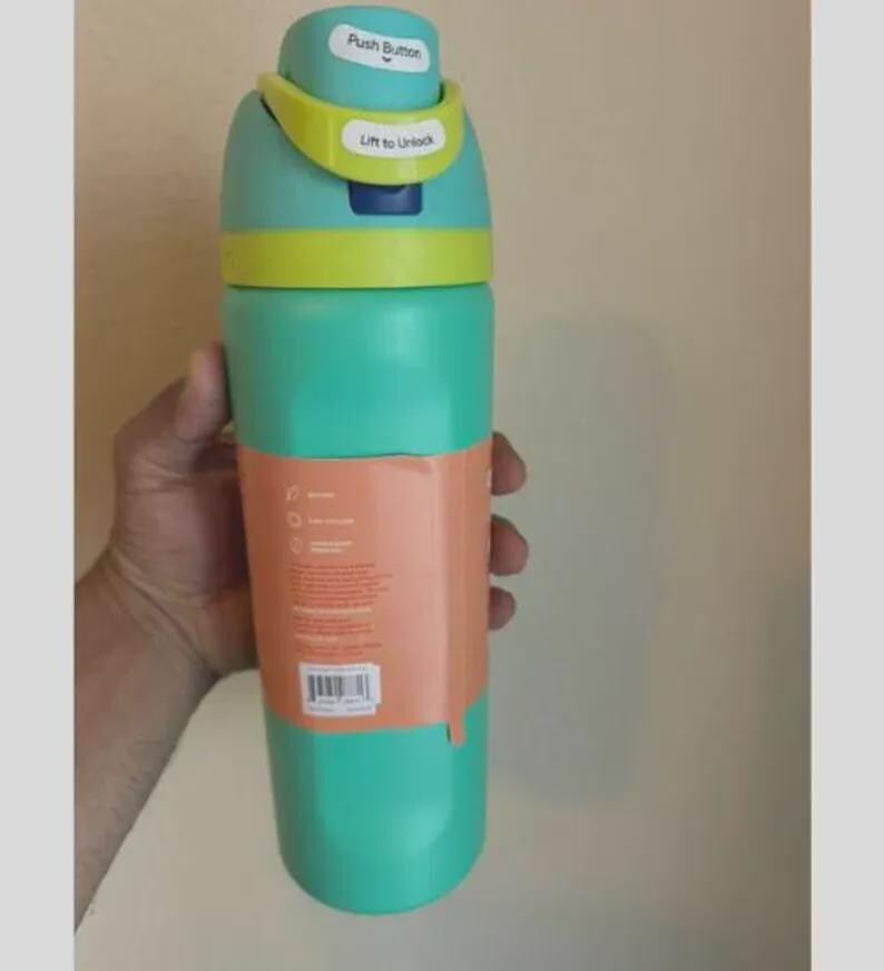 Op de afbeelding: Een talingkleurige waterfles met een limoengroen en talingkleurig deksel met een drukknop en een ontgrendelingsmechanisme. De fles heeft een perzikkleurig papieren etiket rond het midden met gedrukte tekst, iconen en een streepjescode.