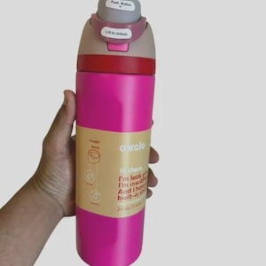 Op de afbeelding: Een felroze Owala ge&iuml;soleerde waterfles met een inhoud van 710 ml. De dop heeft een grijze drukknop en een mauve vergrendeling met het label Lift to Unlock. Een beige papieren band om het midden bevat tekst waaronder Hi there, I'm leak proof.