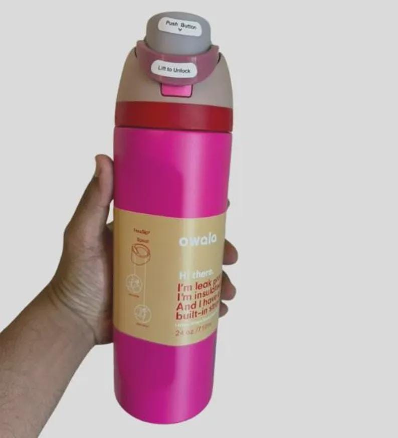 Op de afbeelding: Een felroze Owala ge&iuml;soleerde waterfles met een inhoud van 710 ml. De dop heeft een grijze drukknop en een mauve vergrendeling met het label Lift to Unlock. Een beige papieren band om het midden bevat tekst waaronder Hi there, I'm leak proof.