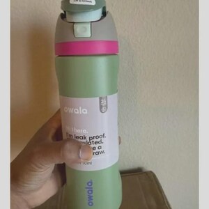 Op de afbeelding: Een saliegroene Owala-waterfles met een grijze en lichtblauwe dop met een roze accentband. De fles heeft een wit label met de tekst Hi there. I'm leak proof. Insulated. I have a straw. De merknaam Owala is in het paars op de fles gedrukt.