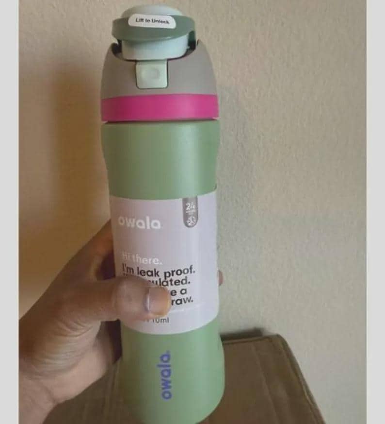 Op de afbeelding: Een saliegroene Owala-waterfles met een grijze en lichtblauwe dop met een roze accentband. De fles heeft een wit label met de tekst Hi there. I'm leak proof. Insulated. I have a straw. De merknaam Owala is in het paars op de fles gedrukt.