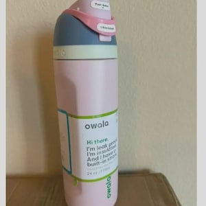 Op de afbeelding: Een lichtroze Owala-waterfles met een blauw en lichtroze deksel met een drukknop en een vergrendeling. Een label op de fles leest: Owala, Hi there. I'm leak proof. I'm insulated. And I have a built-in straw. 710 ml.