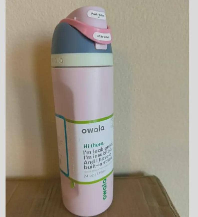 Op de afbeelding: Een lichtroze Owala-waterfles met een blauw en lichtroze deksel met een drukknop en een vergrendeling. Een label op de fles leest: Owala, Hi there. I'm leak proof. I'm insulated. And I have a built-in straw. 710 ml.