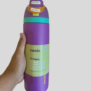 Op de afbeelding: Een paarse Owala-waterfles met een turquoise band, een oranje vergrendelingsmechanisme en een paars deksel. Het label leest: Hi there. I'm leak proof. I'm insulated. And I have a built-in straw. 24 oz / 710 ml. Het deksel bevat de tekst Push Button en Lift to Unlock.